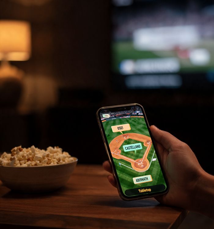 A living room at night: a phone held over a coffee table shows a glowing miniature diamond with name cards while the game plays on the TV.